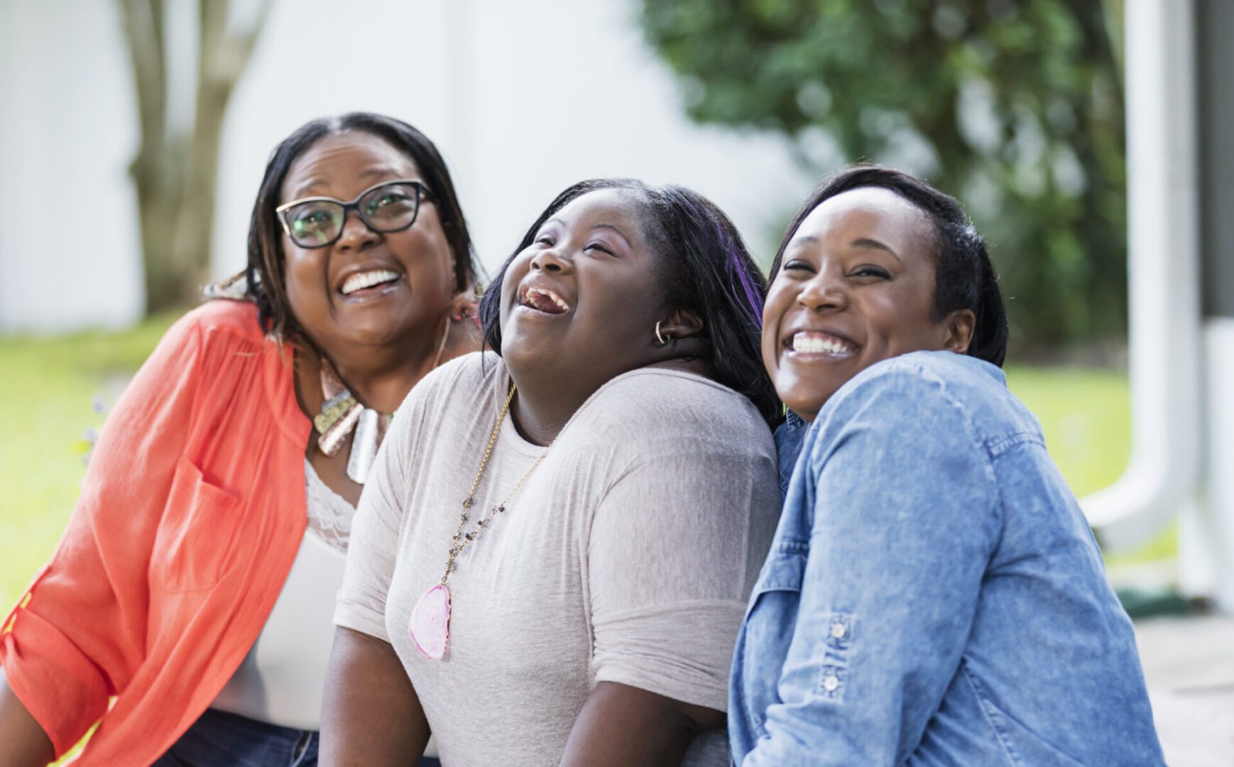 A multi-generation African-American family with grandmother in her 50s, mother in her 30s and 16 year old daughter. The teenage girl, who has down syndrome, is sitting in the middle, looking upward and laughing. Her mother is grinning proudly at the camera.